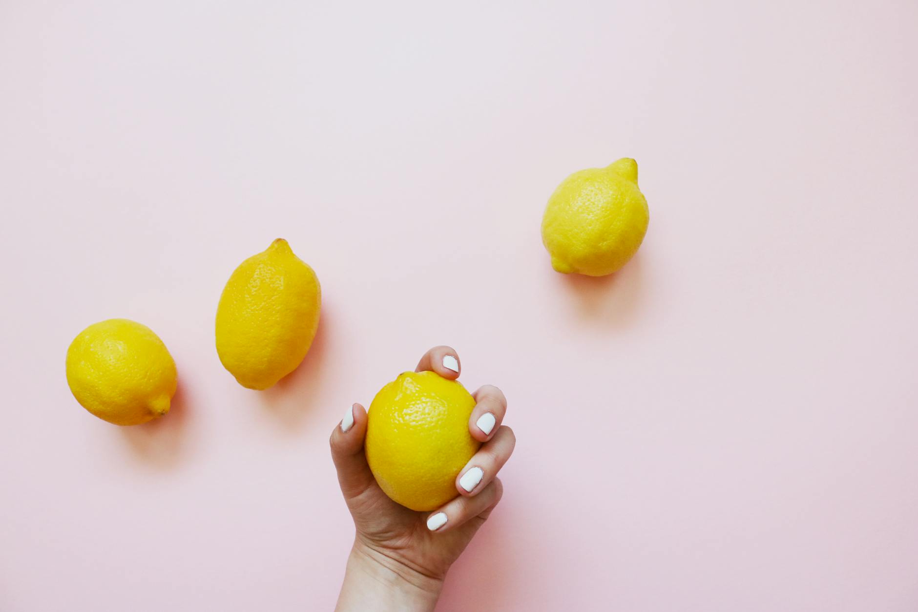 A hand holding a lemon on a soft pink background surrounded by additional lemons